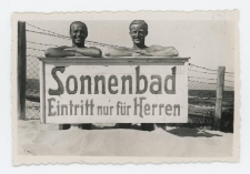 Fotografia czarno-biała przedstawiająca dw&oacute;ch mężczyzn naplaży opartych o tablicę z napisem: &bdquo;Sonnenbad. Eintritt nur f&uuml;r Herren"